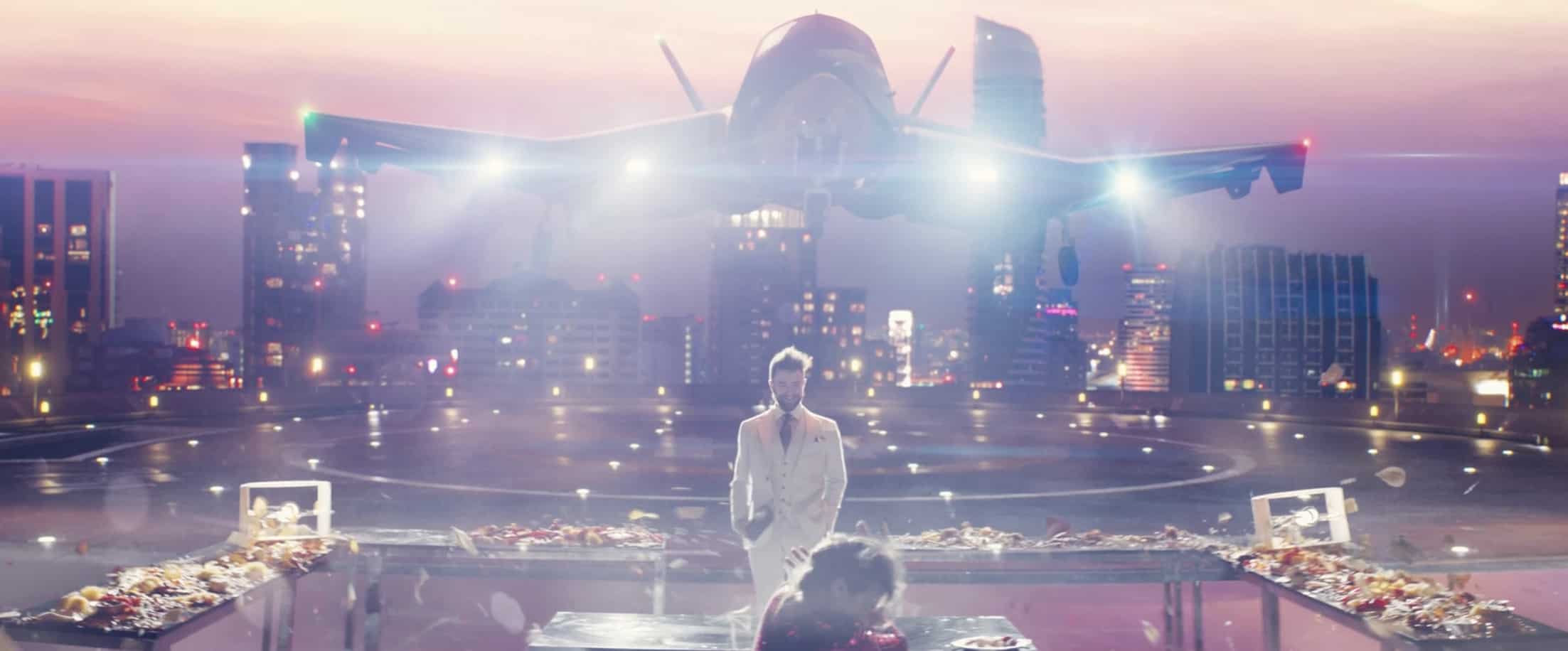 A man in a white suit standing in front of a table with an airplane and city skyline as the backdrop in this image from Amazon Prime Video