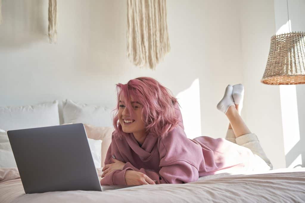 Happy hipster gen z teen girl with pink hair wear hoodie watching tv movie on laptop lying in bed. Smiling teenager make video conference call online, learning online video course, relaxing in bedroom