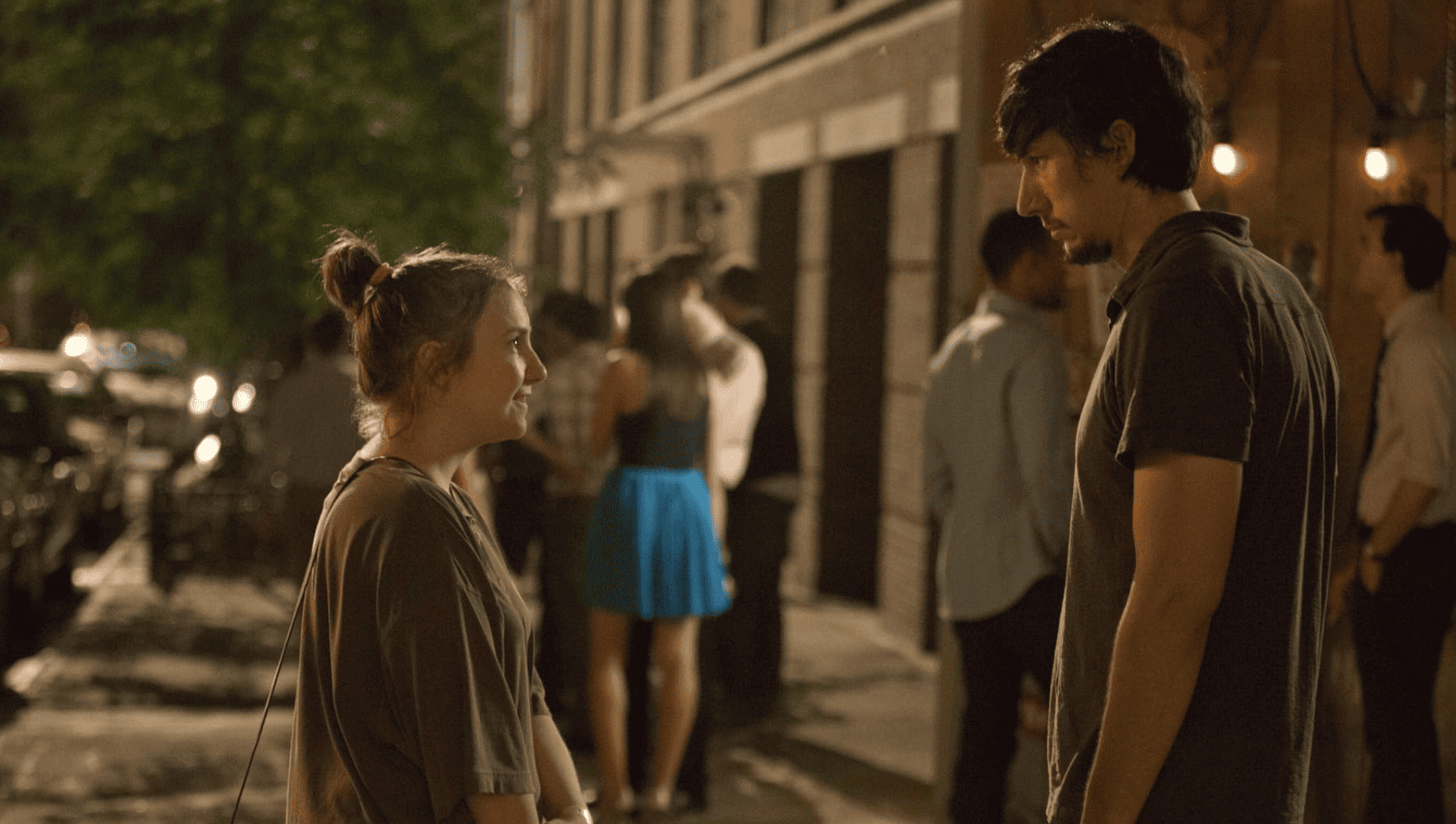 Adam Driver and Lena Dunham standing in the middle of a sidewalk staring at each other while having an intense conversation in this image from Apatow Productions